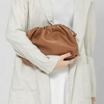 Een vrouw in een lichtbeige blazer en witte jurk houdt de Serena Zachte Leren Pouch Tas van Liora The Label - een minimalistische, moderne, soepele tas - voor haar bovenlichaam. Haar donkere schouderlange haar en zilveren ring maken haar look compleet.