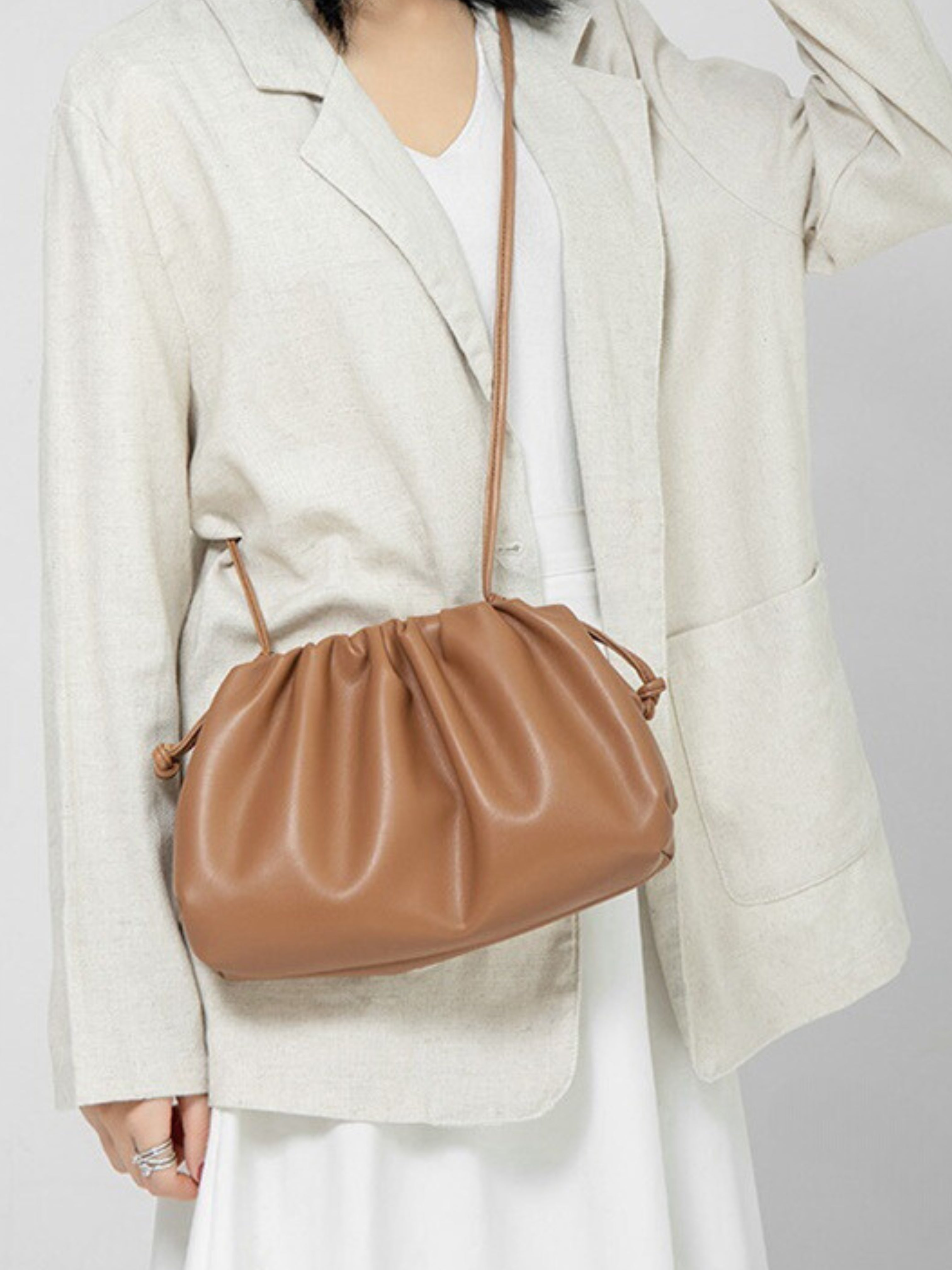 Een persoon in een lichtbeige blazer en witte jurk draagt de Serena Zachte Leren Pouch Tas van Liora The Label, met een minimalistisch, modern ontwerp met soepele leer en een lange riem. De nadruk ligt op de eenvoud van de outfit en het elegante ontwerp van de tas.