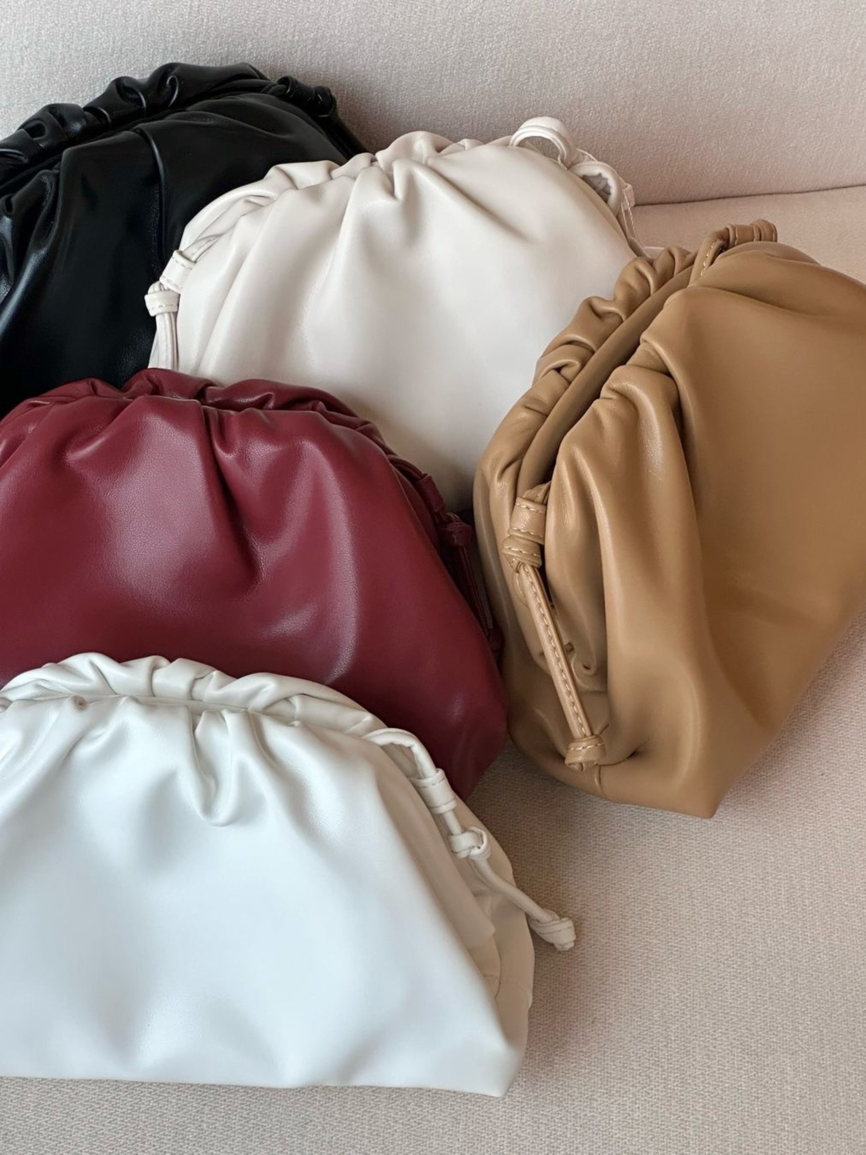Vijf Serena Zachte Leren Pouch Tassen van Liora The Label, in zwart, wit, tan, rood en gebroken wit boterzacht leer, zijn gerangschikt op beige stof. Deze minimalistische, moderne buidels hebben een zacht ruchedesign en overlappen elkaar.