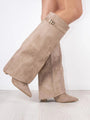 Een persoon staat op een lichthouten vloer tegen een effen witte muur en draagt Liora The Label Dames Kniehoog Laarzen met Brede Flare Bovenkant en Blokhak, met een oversized beige suède look met brede, uitlopende schachten en spitse tenen.
