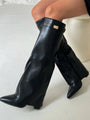 Een persoon draagt Liora The Label Dames Kniehoog Laarzen met Brede Flare Bovenkant en Blokhak in zwart met een goudkleurige gesp. Deze laarzen hebben een spitse neus en gestructureerde wijde pijpen voor een elegante look.