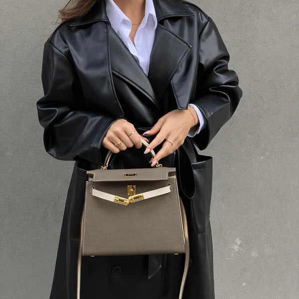 Een persoon in een zwarte leren trenchcoat en wit overhemd houdt de Jovienne Luxe Teal & Tan Leren Handtas van Liora The Label vast, met gouden hardware en een witte riem, staande tegen een effen grijze muur.
