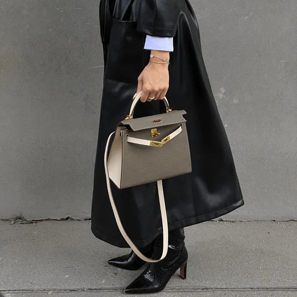 Een persoon in een zwarte leren jas en zwarte laarzen met hak houdt de Jovienne Luxe Teal & Tan Leren Handtas van Liora The Label, met gouden hardware en een witte riem, vast terwijl hij op een betonnen stoep tegen een grijze muur staat.