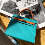 De Jovienne Luxe Teal & Tan Leren Handtas van Liora The Label rust op een houten tafel met een open tijdschrift erachter, zonlicht werpt elegante schaduwen over haar iconische elegantie en tijdloos ontwerp.