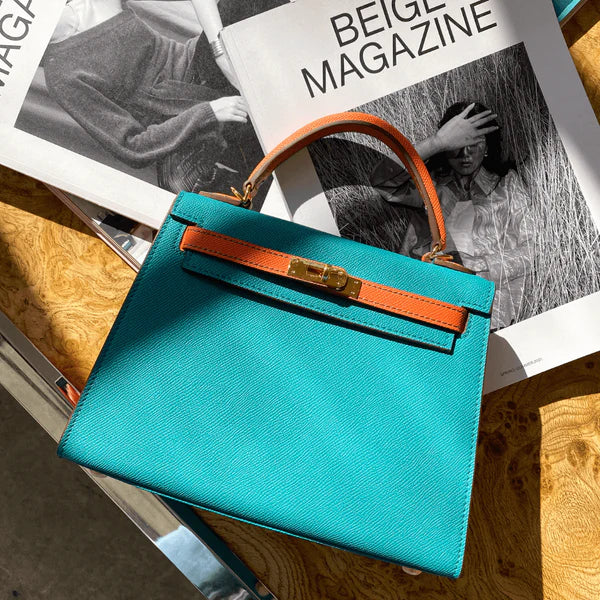 De Jovienne Luxe Teal & Tan Leren Handtas van Liora The Label rust op een houten tafel met een open tijdschrift erachter, zonlicht werpt elegante schaduwen over haar iconische elegantie en tijdloos ontwerp.