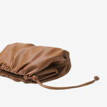 De Serena Zachte Leren Pouch Tas van Liora The Label is een minimalistische, moderne bruine clutch van boterzacht leer met een geplooid ontwerp en trekkoordsluiting, stijlvol gepresenteerd op een witte achtergrond.