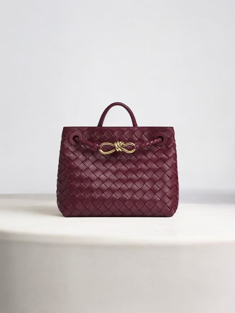 De Alyn Zwarte Geweven Midi Leren Tote Bag van Liora The Label heeft een ontwerp van geweven leer, een handvat aan de bovenkant en een gouden gesp, luxe weergegeven op een wit oppervlak met een effen lichte achtergrond.