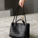 Een hand houdt de Alyn Zwarte Geweven Midi Leren Tote Bag van Liora The Label vast - een elegante tas met gevlochten hengsels - rustend op grijze steen. De persoon draagt beige lange mouwen en een ring, wat moeiteloze stijl en tijdloze aantrekkingskracht benadrukt.