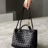 Een hand houdt de Alyn Zwarte Geweven Midi Leren Tote Bag van Liora The Label vast - een elegante tas met gevlochten hengsels - rustend op grijze steen. De persoon draagt beige lange mouwen en een ring, wat moeiteloze stijl en tijdloze aantrekkingskracht benadrukt.
