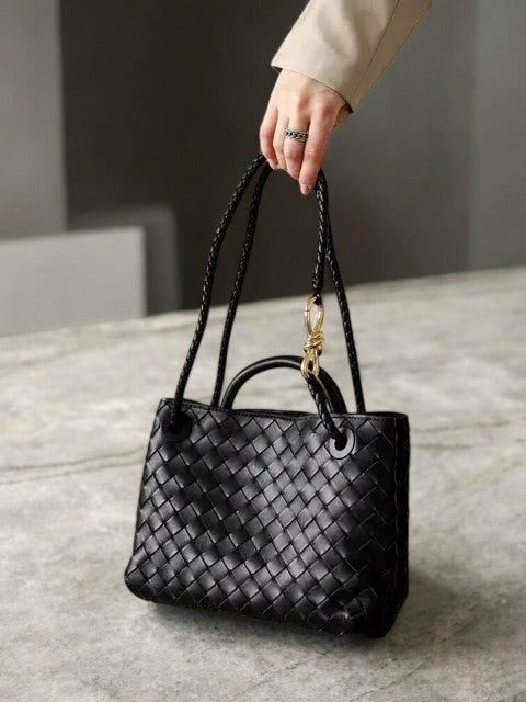Een hand houdt de Alyn Zwarte Geweven Midi Leren Tote Bag van Liora The Label vast - een elegante tas met gevlochten hengsels - rustend op grijze steen. De persoon draagt beige lange mouwen en een ring, wat moeiteloze stijl en tijdloze aantrekkingskracht benadrukt.