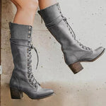 Een persoon met Liora The Label Dames Kniehoge Laarzen met Vetersluiting en Chunky Hak staat bij een lichte muur. De hoge, grijze laarzen hebben decoratieve stiksels en metalen oogjes.