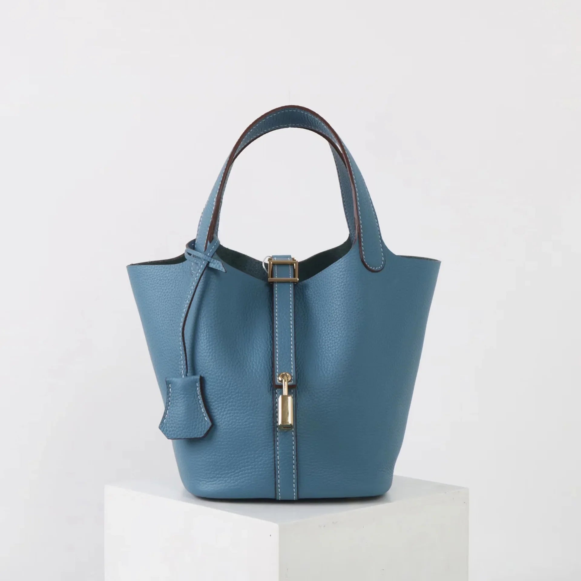 De Elaris Luxe Taupe Leren Emmer Tas van Liora The Label heeft korte hengsels, een decoratieve riem en een goudkleurige gesp. De tas is gemaakt van eersteklas leer en staat op een witte sokkel tegen een effen achtergrond.