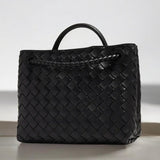 De Alyn Zwarte Geweven Midi Leren Tote Bag van Liora The Label heeft een gestructureerde, rechthoekige vorm en een mandachtige zwartleren textuur, met korte hengsels, elegant weergegeven op een licht oppervlak tegen een neutrale achtergrond.
