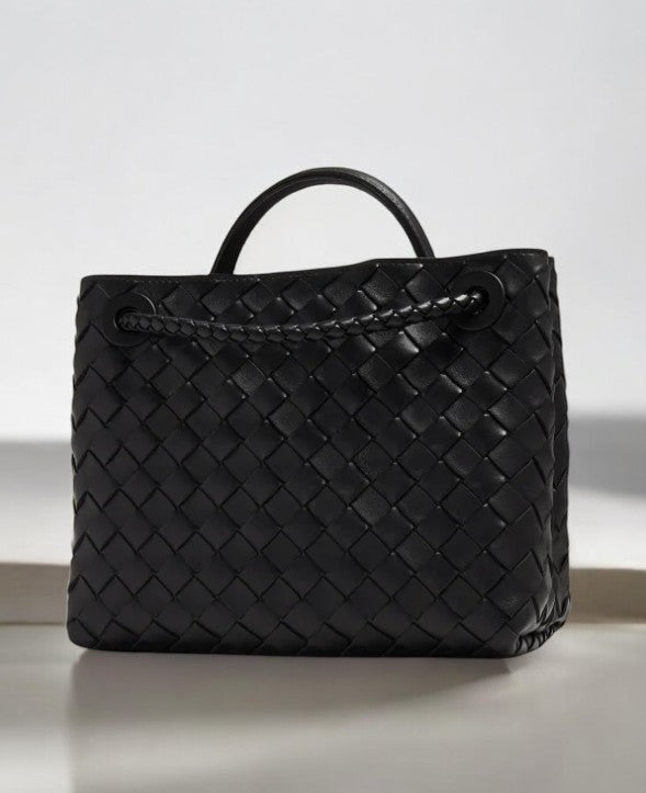 De Alyn Zwarte Geweven Midi Leren Tote Bag van Liora The Label heeft een gestructureerde, rechthoekige vorm en een mandachtige zwartleren textuur, met korte hengsels, elegant weergegeven op een licht oppervlak tegen een neutrale achtergrond.