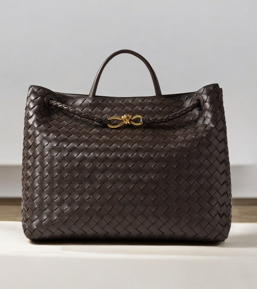 De Alyn Zwarte Geweven Midi Leren Tote Bag van Liora The Label heeft een enkel handvat aan de bovenkant, een gevlochten leren detail aan de voorkant en een gouden knoop accent, weergegeven op een licht oppervlak met een neutrale achtergrond voor tijdloze elegantie.