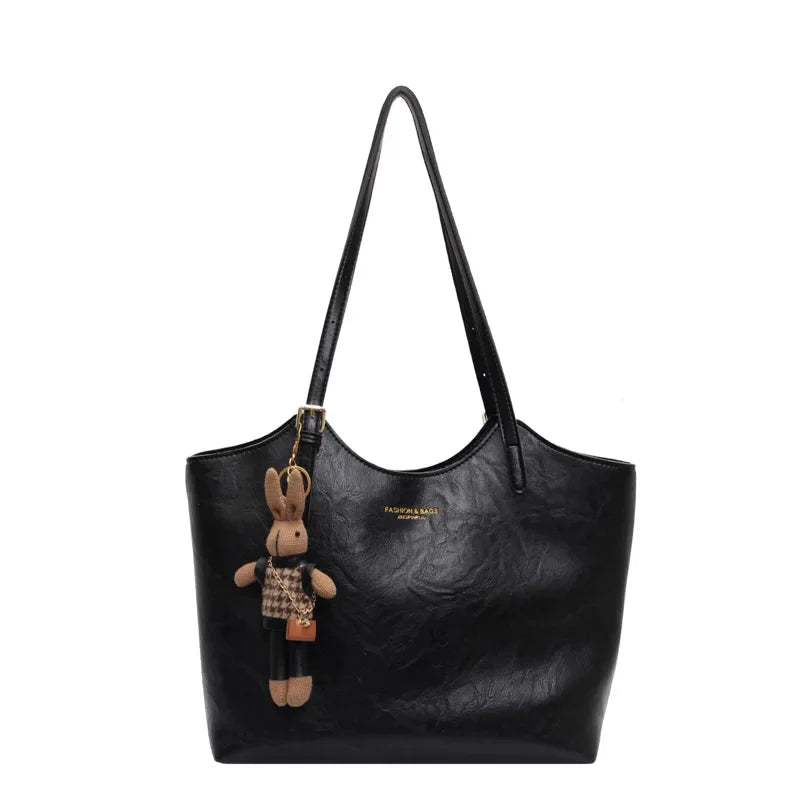 De Thalessa | Chic Leren Tote Bag met Speelse Charme van Liora The Label is een zwarte leren tas met lange riemen, een pluche konijnen sleutelhanger in plaid en elegante gouden branding op de voorkant.