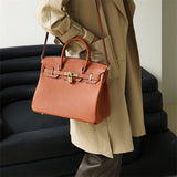 Een persoon in een beige trenchcoat houdt de Isla | Tan Textured Leather Handbag - Klassiek & Tijdloos van Liora The Label vast, een grote tas van structuurleer met goudkleurige hardware. Op de achtergrond staat een moderne zwarte bank.