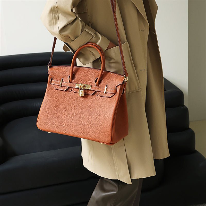 Een persoon in een beige trenchcoat houdt de Isla | Tan Textured Leather Handbag - Klassiek & Tijdloos van Liora The Label vast, een grote tas van structuurleer met goudkleurige hardware. Op de achtergrond staat een moderne zwarte bank.