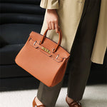 Een persoon in een beige jas en donkere broek houdt binnenshuis de Isla Tan Textured Leather Handtas van Liora The Label vast met twee handvatten en goudkleurige hardware.