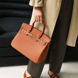Een persoon in een beige jas en donkere broek houdt binnenshuis de Isla Tan Textured Leather Handtas van Liora The Label vast met twee handvatten en goudkleurige hardware.