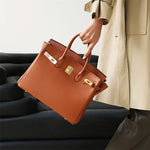 Een persoon in een beige jas en donkere broek houdt de Isla Tan Textured Leather Handbag van Liora The Label vast, met gouden hardware en een slot, terwijl hij bij een zwarte bank staat.