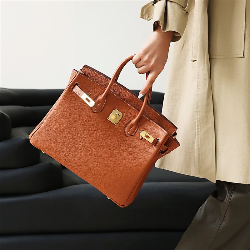 Een persoon in een beige jas en donkere broek houdt de Isla Tan Textured Leather Handbag van Liora The Label vast, met gouden hardware en een slot, terwijl hij bij een zwarte bank staat.