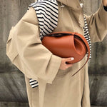 Tegen een betonnen muur houdt een persoon in een beige trenchcoat met een zwart-wit gestreept overhemd de Guinevere Bruine Leren Schoudertas van Liora The Label vast, die moderne elegantie en artistieke raffinement belichaamt.