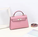 De Aurora Signature Mini Leren Handtas van Liora The Label is een roze, gestructureerde tas met bovenhandvat in volnerf leer met gouden hardware en gespsluiting. De tas heeft een afneembare riem en rust op een wit vlak bij een wit fotolijstje.