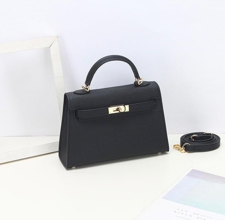 De Aurora Signature Mini Leren Handtas van Liora The Label - een zwarte, gestructureerde tas met bovenhandgreep, gouden hardware en een afneembare schouderriem - rust op een wit oppervlak naast een lege lijst en een pastelkleurig blad.
