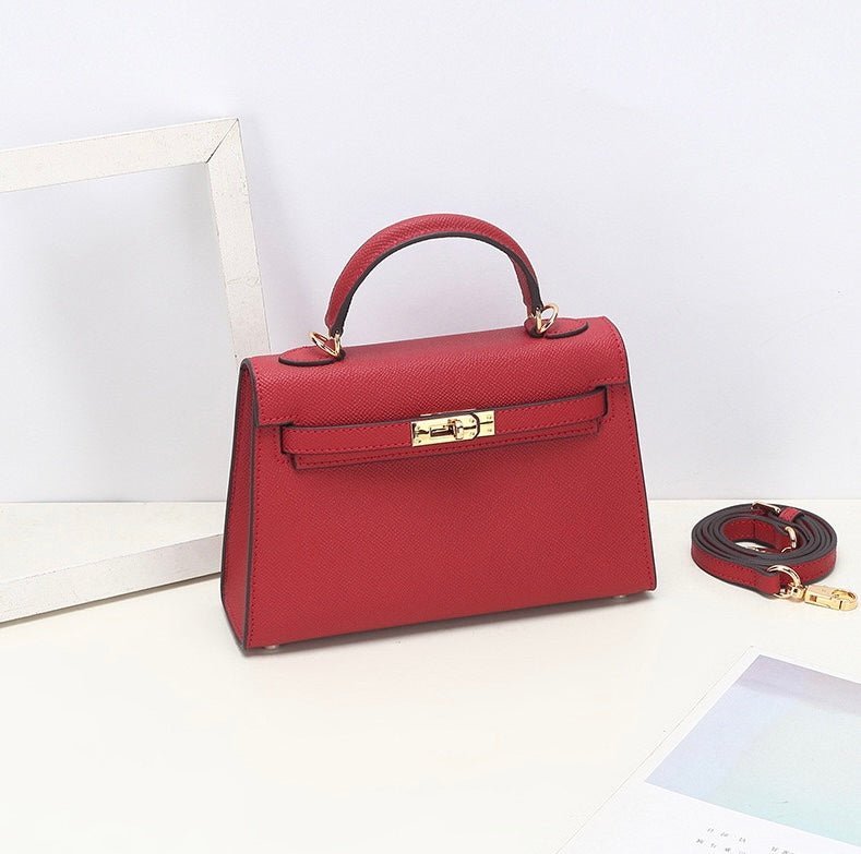 De Aurora Signature Mini Leren Handtas van Liora The Label is een gestructureerde top handtas van volnerf leer met een gouden sluiting en afneembare schouderriem, gepresenteerd op een witte ondergrond naast een wit frame en deels zichtbaar papier.
