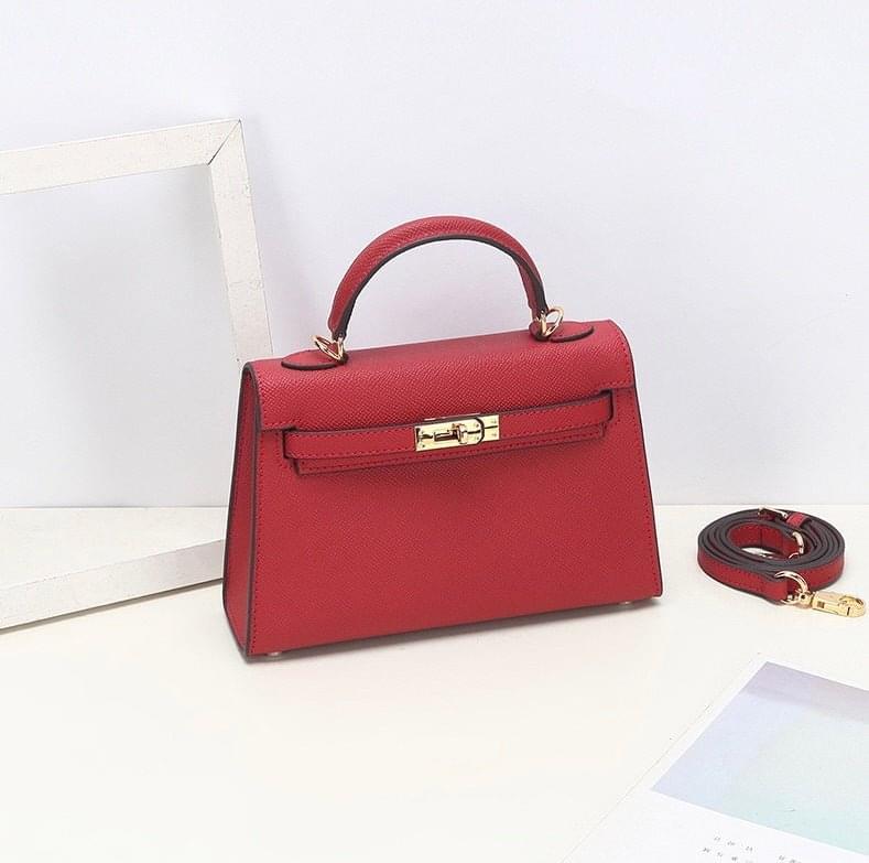 De Aurora Signature Mini Leren Handtas van Liora The Label is een gestructureerde top handtas van volnerf leer met een gouden sluiting en afneembare schouderriem, gepresenteerd op een witte ondergrond naast een wit frame en deels zichtbaar papier.