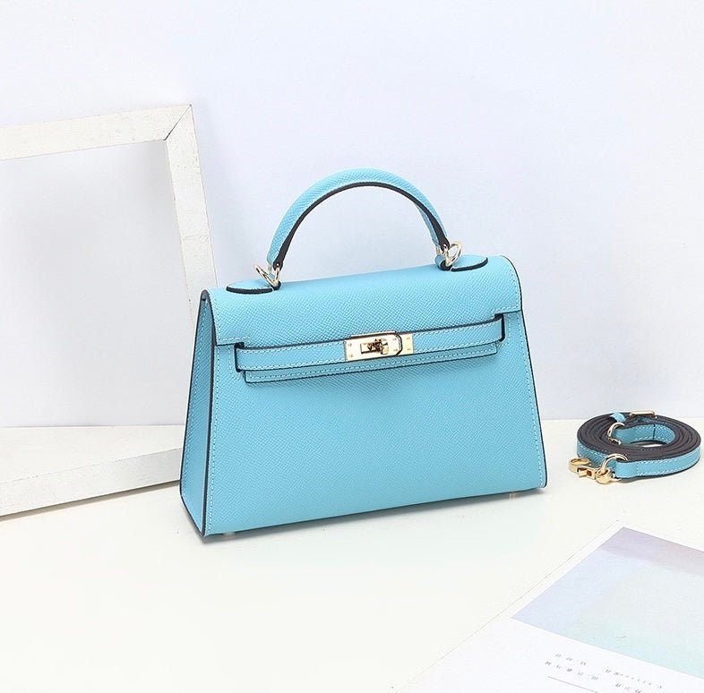 De Aurora Signature Mini Leren Handtas van Liora The Label-lichtblauw, gestructureerd, gemaakt van volnerf leer met gouden hardware-staat op een witte ondergrond met een afneembare riem, witte fotolijst en kalender. De achtergrond is lichtblauw.