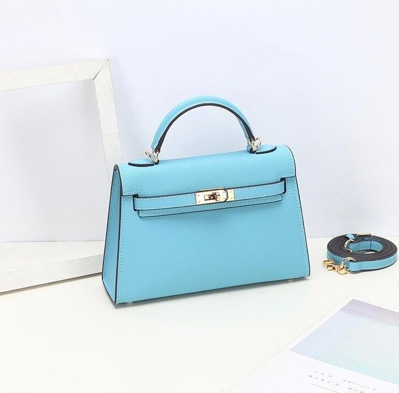 De Aurora Signature Mini Leren Handtas van Liora The Label-lichtblauw, gestructureerd, gemaakt van volnerf leer met gouden hardware-staat op een witte ondergrond met een afneembare riem, witte fotolijst en kalender. De achtergrond is lichtblauw.