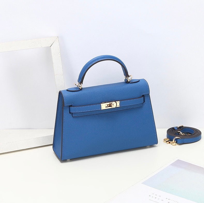 De Aurora Signature Mini Leren Handtas van Liora The Label, een gestructureerde blauwe minitas in volnerf leer met een bovenhandvat en gouden hardware, staat op een wit vlak met de bijpassende schouderriem ernaast.