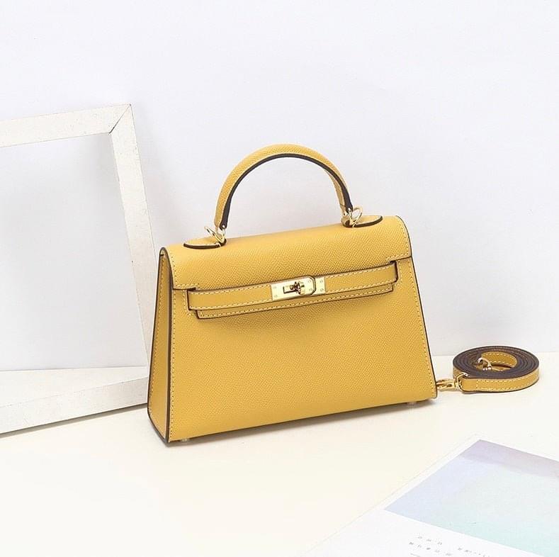 De Aurora Signature Mini Leren Handtas van Liora The Label in geel volnerf leer, met top handvat, gouden sluiting en afneembare schouderriem, wordt op een witte ondergrond bij een lege witte fotolijst getoond.