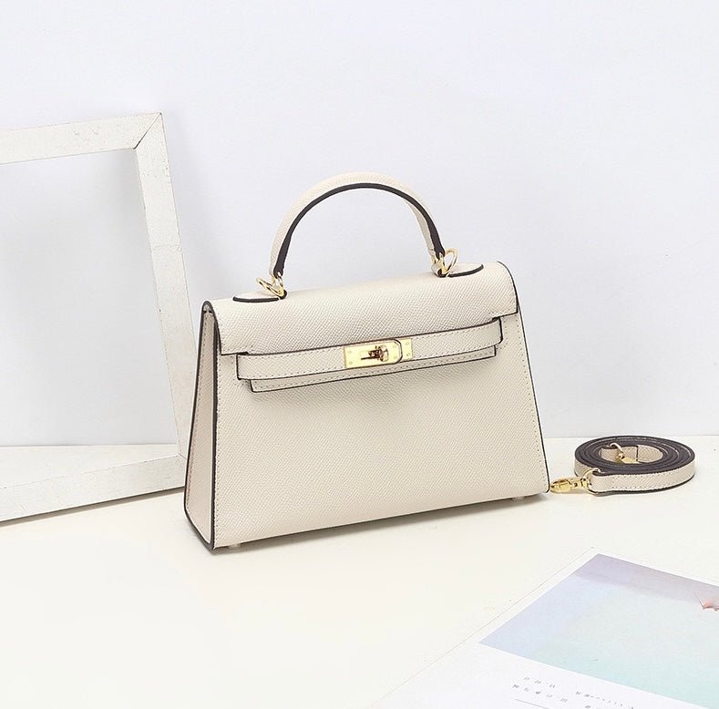 De Aurora Signature Mini Leren Handtas van Liora The Label, in crème volnerf leer met een bovenhandgreep, gouden hardware en een afneembare schouderriem, wordt tentoongesteld op een wit oppervlak naast een fotolijstje en een tijdschrift.