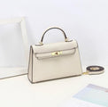 De Aurora Signature Mini Leren Handtas van Liora The Label, in crème volnerf leer met een bovenhandgreep, gouden hardware en een afneembare schouderriem, wordt tentoongesteld op een wit oppervlak naast een fotolijstje en een tijdschrift.
