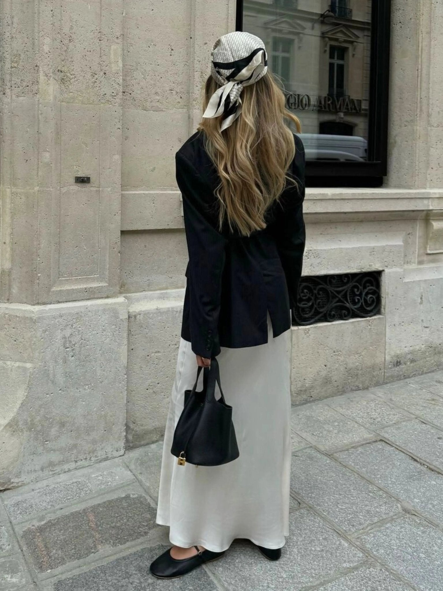 Een vrouw met lang, golvend blond haar staat op een stenen stoep, met haar gezicht naar voren, en houdt de Elaris Luxe Taupe Leren Emmer Tas van Liora The Label vast - een elegante, tijdloze tas - in een stedelijke omgeving. Ze draagt een sjaal met patroon en een zwarte blazer.