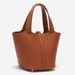 De Elaris Luxe Taupe Leren Emmer Tas van Liora The Label heeft een tijdloos, elegant ontwerp met dubbele handgrepen, goudkleurige pootjes en een gestructureerd minimalistisch silhouet, weergegeven op een effen witte achtergrond.