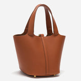 De Elaris Luxe Taupe Leren Emmer Tas van Liora The Label heeft een tijdloos, elegant ontwerp met dubbele handgrepen, goudkleurige pootjes en een gestructureerd minimalistisch silhouet, weergegeven op een effen witte achtergrond.
