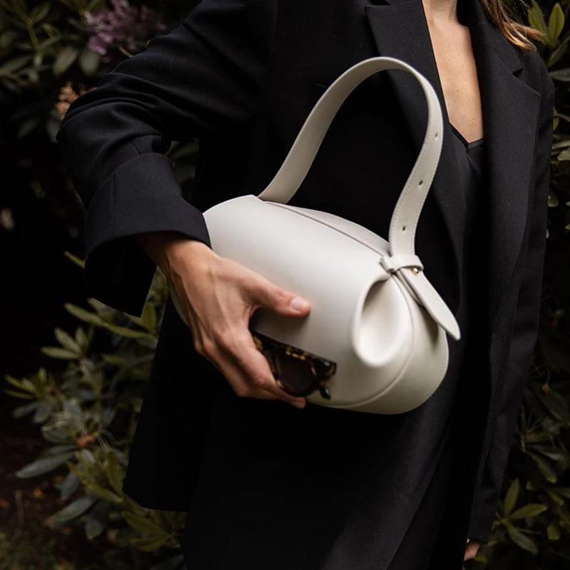 Een persoon in het zwart houdt de Liora The Label Guinevere gesculpteerde bruin lederen schoudertas vast, die moderne elegantie en artistieke verfijning belichaamt. De brede riem en de gehaakte zonnebril maken de look compleet, met bladgroen op de achtergrond.
