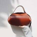 Een hand houdt de Guinevere Sculpted Bruine Leren Schoudertas van Liora The Label vast-moderne elegantie en artistiek raffinement tegen een minimalistische witte achtergrond met zachte schaduwen.