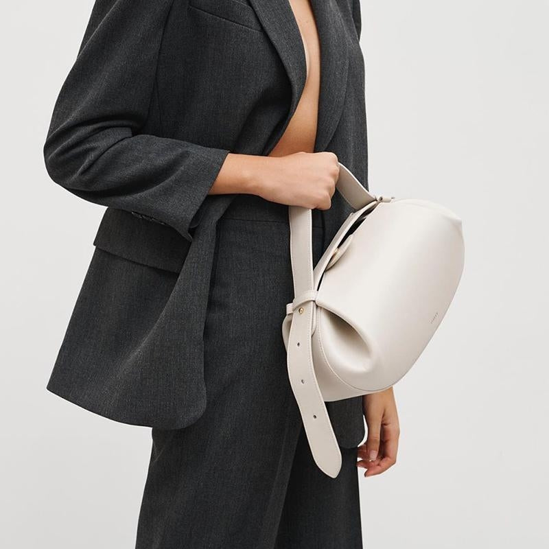 Een persoon in een donkergrijs pak zonder overhemd houdt de Guinevere | Sculpted Bruine Leren Schoudertas van Liora The Label vast, die minimalistische luxe uitstraalt. De effen lichte achtergrond benadrukt de moderne elegantie en artistieke raffinement van de tas.
