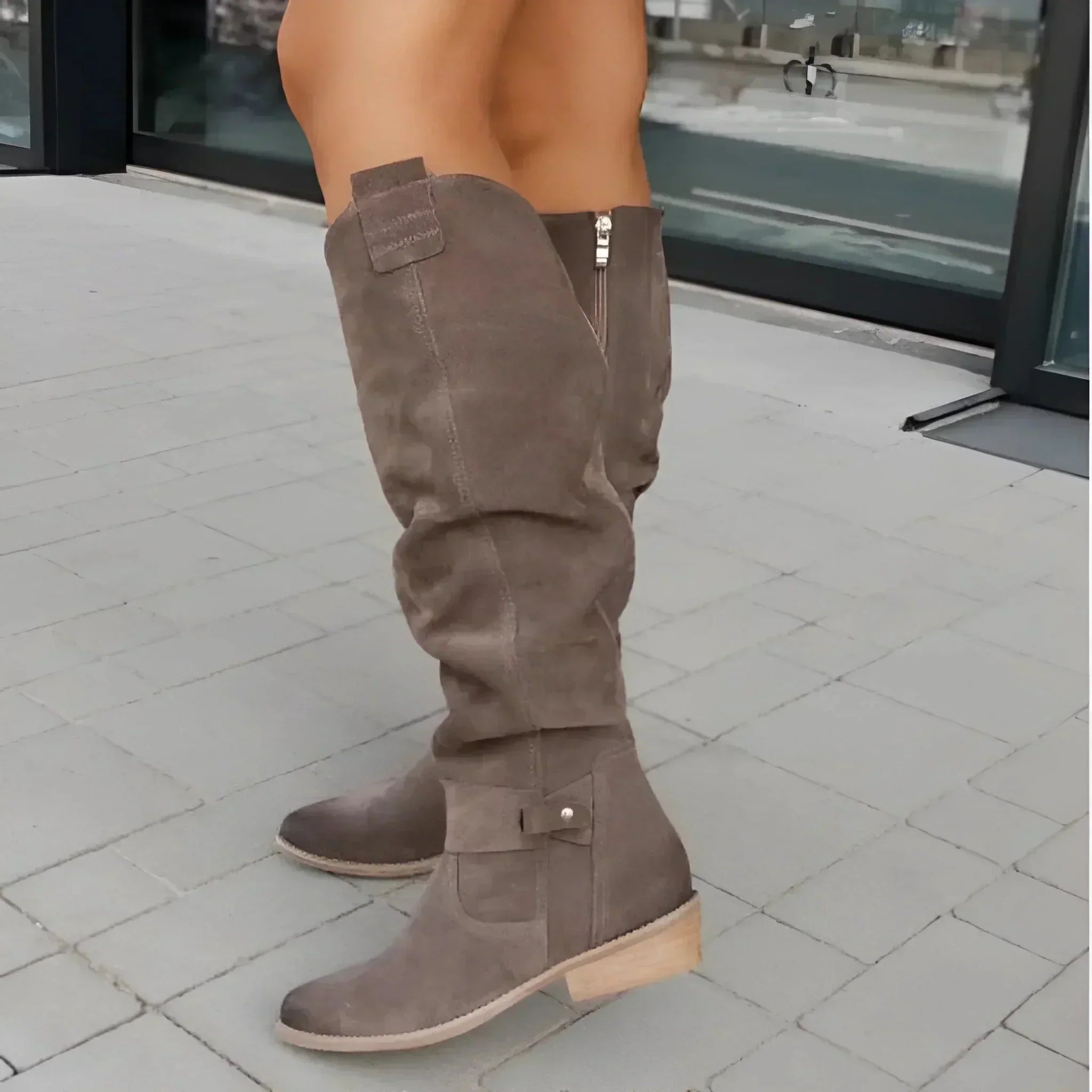 Buiten draagt een persoon Liora The Label Capucine Leren of Suède Boots voor Dames in taupe suède, met een lage houten hak, rits aan de zijkant en tijdloos ontwerp op lichtgrijze tegels.